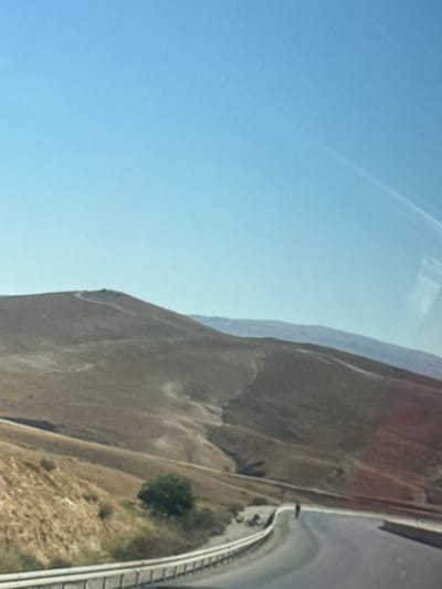The image shows a landscape in al-Qasoum Regional Council, Israel. The scene is dominated by a mountain or hill on the right side, with a predominantly barren and rocky terrain. A road runs alongside the mountain, separated by a barrier. A single tree is visible along the road, along with some sparse vegetation. The sky occupies a significant portion of the left side, appearing clear and blue.