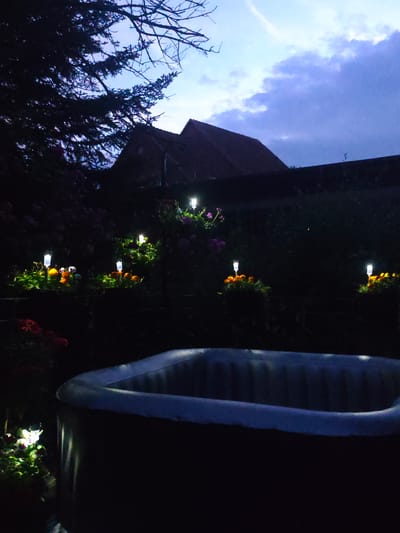 In Lügde, Germany, at dusk, an inflatable hot tub is situated in a dark backyard. The sky above is a deep blue with scattered clouds, transitioning from twilight to night. In the background, the silhouette of a building with a tiled roof is visible. The foreground is dominated by the hot tub, and to its left, solar-powered garden lights illuminate flower beds filled with bright orange and yellow marigolds and some pink flowers. A large, dark evergreen tree is visible in the upper left corner of the frame, with its branches silhouetted against the sky.