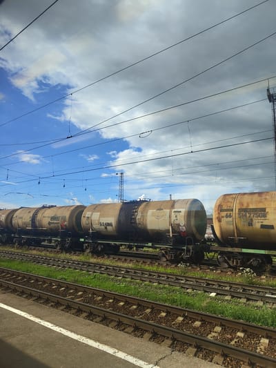 The image depicts a train of tank cars parked on railroad tracks under a cloudy sky. The train cars are cylindrical and appear to be made of metal, with some showing signs of rust and wear. They are predominantly light brown or beige in color, with some lettering and numbers visible on their sides. The train is positioned on multiple sets of tracks, with grass and ballast visible between them. In the background, power lines and poles crisscross the sky, and a tall lattice structure, possibly a telecommunications tower or part of the railway infrastructure, is visible on the right. The overall impression is one of an industrial or logistical setting, typical of a railway yard. The location is identified as Фруктовая, Russia. There are no people or animals visible in the image. The scene is static, with the train at rest.