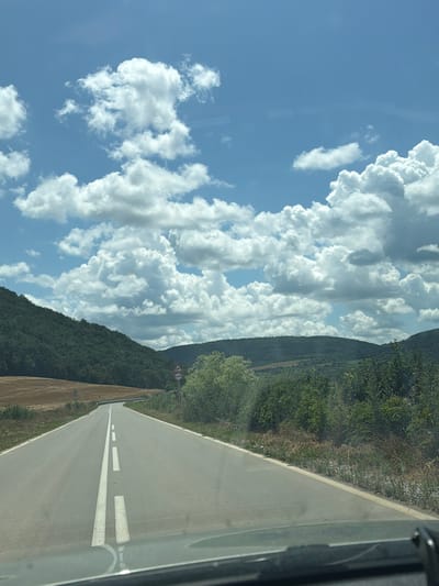 The image depicts a scenic view from a vehicle traveling on a road. The road itself is paved and has white lane markings, indicating it is a well-maintained route. To the left of the road, a steep, tree-covered hillside rises, with lush green foliage dominating the landscape. In the sky, large, fluffy white clouds are scattered across a bright blue backdrop, suggesting a clear and pleasant day. The time of day appears to be daytime, likely late morning or afternoon, given the brightness of the sky and the shadows cast.

The perspective suggests the photo was taken from the passenger side of a car, looking forward and slightly to the left. A small, circular road sign with two arrows pointing in opposite directions is visible on the side of the road, indicating a hazard or upcoming change in road conditions. The overall mood is peaceful and suggests a journey through a rural or mountainous area. The location is identified as Garchinovo, Bulgaria. There is no visible text other than the road sign.