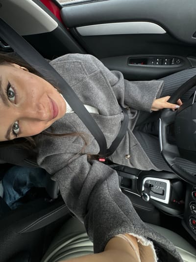 A woman is taking a selfie from the driver's seat of a car. She is wearing a gray coat and pinstripe pants, and a seatbelt is fastened. The car's interior is visible, including the steering wheel, dashboard, gear shift, and door controls. The exact location in Dubki (Gp Kubinka), Russia cannot be determined from the image.