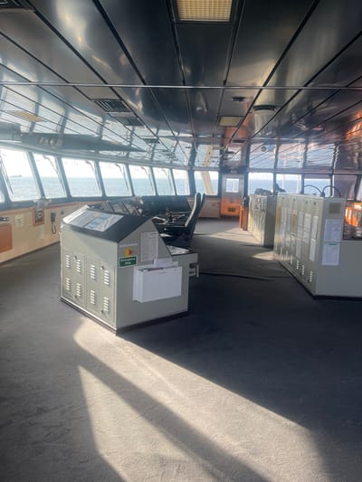 The image displays the unoccupied bridge of a ship, located off the coast of Orimedu, Nigeria. The interior features a dark grey carpeted floor and a reflective, segmented dark grey ceiling containing recessed lighting fixtures. A panoramic array of large windows provides an unobstructed view of a calm blue sea under a bright sky, with a distant ship visible on the horizon to the left.
In the left foreground, a light grey console unit stands with louvered panels, a dark top surface holding various documents, a green "Emergency Stop" sign, and a white wall-mounted document holder attached to its front. Further into the midground, a primary ship's control console equipped with multiple screens, levers, and a single black captain's chair is positioned facing the windows. To the right, two light grey utility cabinets are present, each featuring multiple framed documents or labels on their fronts. No individuals are visible. Bright natural light enters through the windows, creating pronounced linear shadows across the carpet.