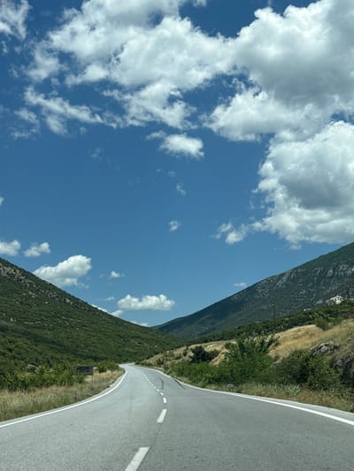 This is a point-of-view shot from a vehicle traveling on a paved road that curves through a mountainous landscape under a blue sky dotted with white clouds. The road has a white broken line separating lanes and solid white lines along the edges. To the left, a densely vegetated, green hillside rises steeply. To the right, a drier, more arid hillside with sparse vegetation and rocky outcrops is visible. In the distance, more mountains are visible, receding into the haze. A signpost with some text is visible on the left side of the road. The environment is natural and appears to be rural. The exact location within the Prosotsani Municipal Unit, Greece, is not discernible from the image alone.