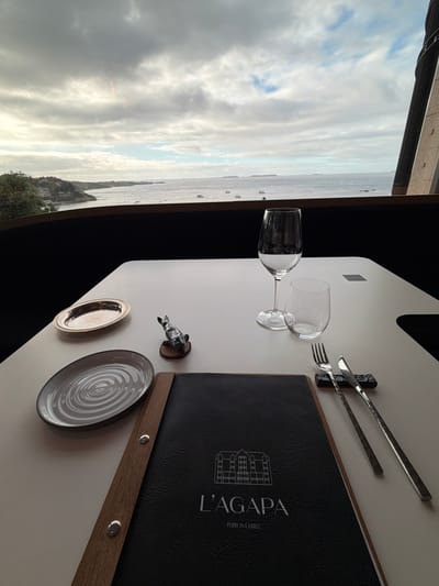 This image captures a table setting for a meal with a scenic view of the sea and a cloudy sky. The table is set with two wine glasses, one filled with a dark liquid, a fork and knife resting on a dark holder, and a menu with the text "L'AGAPA" and "PERROS-GUIREC" visible. There's also a small silver dish and a decorative metal figurine of an animal, possibly a dog, on the table.
The background reveals a panoramic view of the sea, with islands or landmasses visible in the distance, along with several boats. The sky is filled with dramatic clouds, suggesting it might be overcast or that a storm is approaching. The overall lighting implies it is daytime, possibly late afternoon, given the diffused light. The presence of the menu and table setting suggests an ongoing or imminent dining experience. The setting appears to be a restaurant or dining area with a large window offering the sea view.