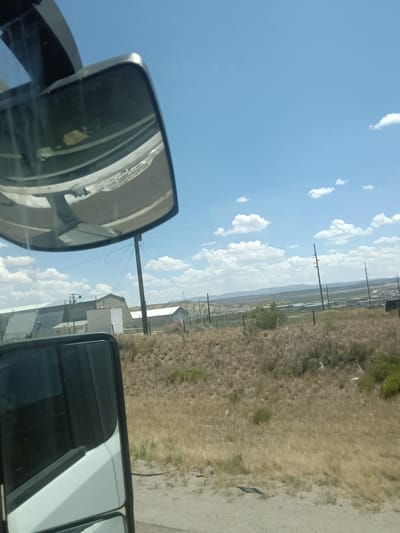 The image depicts a rural landscape with industrial elements under a bright blue sky with scattered white clouds. In the foreground, a side-view mirror of a vehicle reflects a portion of the sky and what appears to be the vehicle's interior, possibly showing the dashboard and windshield. To the left, a white and black vehicle is partially visible.

The middle ground features a collection of industrial buildings, some with large, flat roofs and metallic siding. One building has a stone facade. A tall wooden utility pole stands prominently in the center of the image, with power lines stretching across the scene. Dry, golden-brown grass and sparse green bushes cover the ground, indicative of an arid environment.

In the background, a range of distant hills or mountains is visible, and more utility poles are scattered throughout the landscape. The overall impression is that of an industrial area situated in a semi-arid, open region. The location is identified as Purple Sage, United States of America, which suggests this is a view of the outskirts or an industrial zone within or near that area. No people are visible in the image.