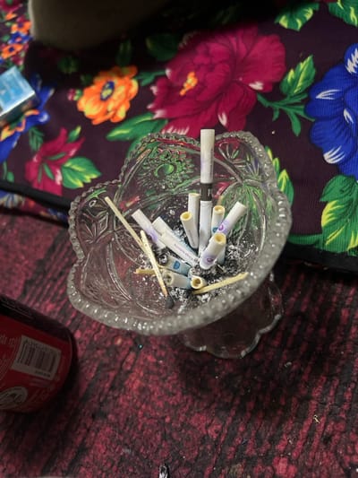 A crystal ashtray overflows with Vegas Nights cigarette butts and spent matches. The ashtray rests on a surface adorned with a vibrant floral pattern. To the right, a partially visible red Coca-Cola can sits on a worn, dark red surface with a rough texture. The composition suggests a scene of casual disarray and consumption.