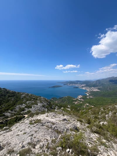The image captures a high-angle view along the coast of Vrijesno, Montenegro. The azure waters of the Adriatic Sea meet the rocky coastline, and a stretch of the town is visible along the shore. A mountain slope, covered with sparse vegetation and exposed rock, dominates the right side of the frame. The sky is a clear blue with scattered, fluffy white clouds on the left.