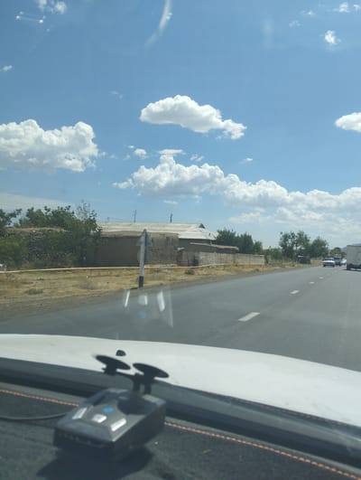 The image depicts a view from inside a vehicle, looking out through the windshield at a road in Asmansay, Uzbekistan. The sky is a clear blue with scattered white cumulus clouds. On the right side of the road, there are several vehicles, including what appears to be a white truck and other cars. The road itself is asphalt with white lane markings. To the left of the road, there is a dirt or gravel embankment with sparse vegetation. Further back, there are low-lying buildings with flat roofs, constructed of mud or adobe. Some trees and bushes are visible in the background, and there are power lines and poles. In the foreground, on the dashboard of the vehicle, a black radar detector with two suction cups is visible. The overall scene suggests a sunny day in a rural or semi-rural area.