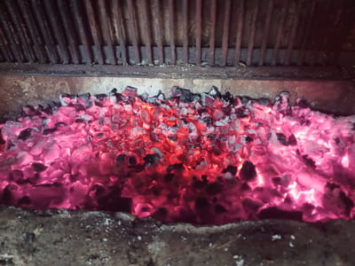 This is a close-up, eye-level, interior shot of a barbecue grill filled with glowing red and pink embers. The embers are densely packed and appear to be wood or charcoal. Above the embers, a metal grate made of thick, vertical rods is visible, showing signs of rust and soot. The background behind the grate is dark and indistinct. The scene is located in Nova Mohylnytsia, Ukraine. The image is well-lit by the heat and light of the embers. There are no people or animals visible in the image.