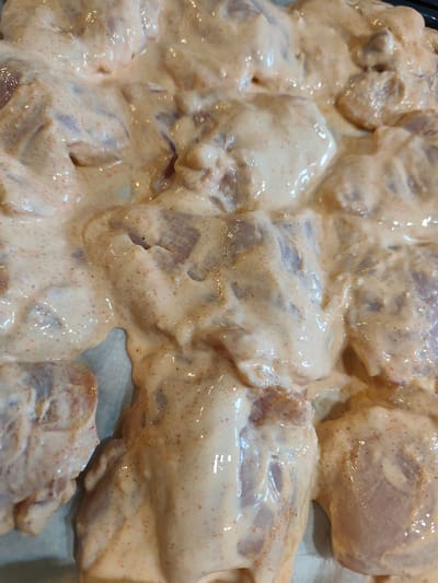 The image shows a close-up, overhead view of raw chicken pieces coated in a creamy, light orange marinade with visible red spice specks. The chicken pieces are arranged in a single layer on a light-colored surface, possibly a baking sheet lined with parchment paper. The marinade appears thick and smooth, with some pieces of chicken showing through the coating. The lighting is bright, creating highlights on the moist surface of the marinade and chicken. There are no people, animals, or external environmental elements visible in the image. The scene depicts food preparation.