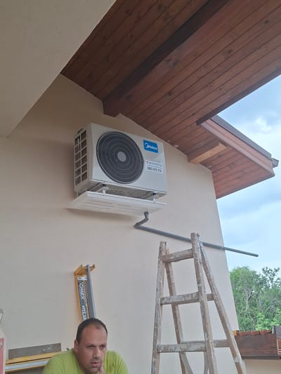 The image depicts the exterior wall of a building in Dolna Malina, Bulgaria, under a wooden-paneled overhang. An outdoor air conditioning unit, a Midea model, is mounted on the wall. Below the unit, a ladder made of weathered wood leans against the wall, and a horizontal pipe extends from the unit across the wall. To the left of the ladder and further down the wall, a man in a lime-green t-shirt is seated, looking downwards, with a tool that has "CHEMEX HTC12" visible on it behind him. The sky is overcast.