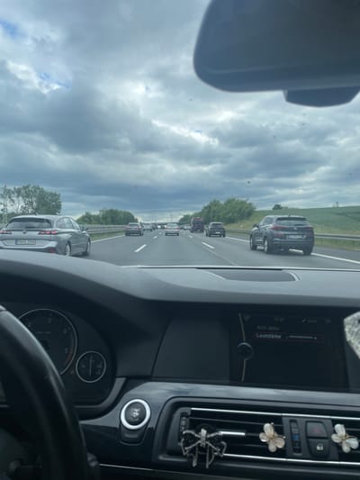 The image depicts a first-person view from inside a vehicle on a multi-lane German Autobahn, situated in or near Staufenberg, Germany. The time displayed on the vehicle's infotainment system is 17:47. The sky is heavily overcast with grey clouds, featuring small patches of visible blue sky.
The perspective is from the driver's seat of what appears to be a BMW, based on the partial view of the steering wheel, the dashboard layout, and the "START STOP ENGINE" button on the central console. The instrument cluster shows a tachometer on the left and a combination temperature/fuel gauge on the right. The infotainment screen displays "AUX aktiv" and "Lautstärke" (volume). Attached to the air vents below the screen are three decorative clips: a black glittery bow on the left and two white and gold butterfly clips on the right. Faint, circular reflections or dust particles are visible on the windshield, particularly against the sky.
The highway is experiencing moderate to heavy traffic flow. In the adjacent left lane, a silver Peugeot 308 SW station wagon is visible, bearing the German license plate "PAN L 2286". Further ahead in the right lane, a dark grey Hyundai Tucson SUV is observed with the German license plate "GO BW 119". Other assorted vehicles, including a red van, are present in the distance across various lanes.
The road surface is asphalt with clear white lane markings. A green median strip separates the opposing directions of traffic, bordered by a metallic guardrail. The surrounding environment consists of green fields and sections of dense trees on both sides of the highway. Overhead, power lines equipped with red and orange spherical marker balls are strung across the landscape.