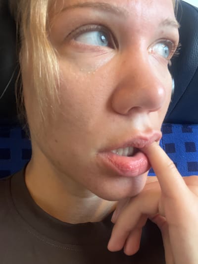 A close-up, low-angle shot shows a person's face and hand. The person, with light skin and blonde hair, is looking up and to the left, with their blue eyes clearly visible. Their mouth is slightly open, with their lips appearing plump and glossy. A finger from their right hand is touching their upper lip, near the corner of their mouth. The background is a mix of dark surfaces, one of which has a blue, checkered pattern, and a brown shirt or fabric. The lighting suggests it could be daytime. The person's expression is difficult to discern definitively due to the close-up crop, but the gesture of touching their lip could suggest contemplation or a playful moment. There is no visible text in the image. The scene is intimate and personal, focusing on the interaction between the hand and the face.