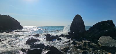The image shows a rocky coastline on a clear, sunny day. The foreground is dominated by dark, wet rocks of various sizes, with waves crashing against them and creating white foam. In the mid-ground, the sea is a vibrant blue, sparkling with sunlight. A large, pointed rock formation stands prominently in the right mid-ground, with a wave breaking against its base. In the background, the sea meets the horizon under a clear, pale blue sky. Two faint white contrails are visible in the sky. The scene depicts a natural coastal landscape.