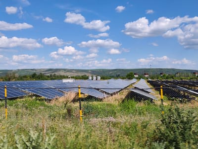 The image depicts a solar farm under a bright, partially cloudy sky. The foreground is filled with wild grass and some flowering plants, with a yellow and black striped pole marking a boundary. Behind the vegetation, rows of blue solar panels are neatly arranged on metal frames, angled to capture sunlight. Some panels are in sharp focus, while others further in the distance are slightly blurred. In the middle ground, a few small white buildings with windows are visible among the panels. The background shows rolling hills covered in green vegetation and trees. In the far distance, there are some industrial-looking structures and light poles. The sun is high in the sky, suggesting it is daytime, likely late morning or early afternoon, given the bright lighting and clear blue sky with scattered white cumulus clouds. There is no visible text in the image. The scene appears to be a rural or semi-rural area, possibly an open field or undeveloped land dedicated to renewable energy generation in Krushovitsa, Bulgaria. The overall mood is one of peaceful productivity, with the solar panels harnessing natural energy under a pleasant weather conditions.