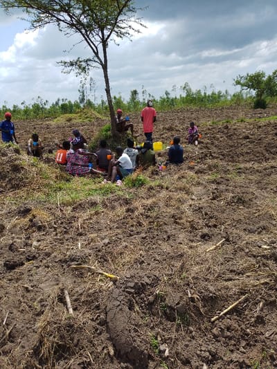 A group of people rests in a field. They sit and stand near a small tree, some holding cups and a yellow bucket. The field is brown and appears to be recently tilled, with dry grass and stalks scattered across the soil. In the background, a line of trees extends to the horizon under a cloudy sky. The composition suggests a break during agricultural work.