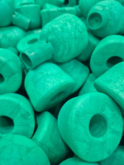 The image is a close-up, vertical shot of a pile of teal-colored, doughnut-shaped objects. The objects are textured and appear to be made of a matte material, possibly plastic or ceramic. They are arranged in a jumbled fashion, with some overlapping and others partially obscured. The lighting creates shadows that highlight the contours of the objects. The exact location in Pabradė, Lithuania, is not discernible from the image itself.