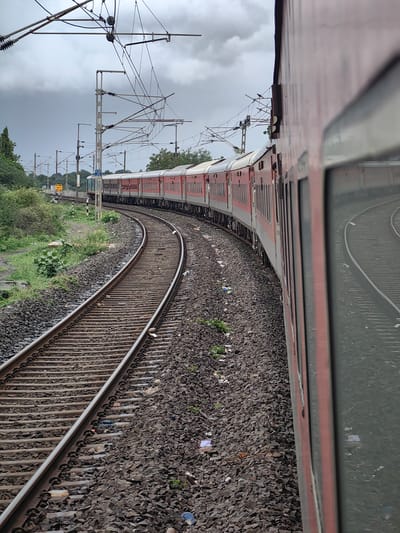 An exterior, eye-level, slightly tilted shot captures a passenger train on a curved track under a cloudy sky. The train, predominantly red and white, is seen from the side, its windows reflecting the surrounding environment. The track, made of gravel and metal rails, curves to the left, disappearing into the distance. Vegetation, including trees and bushes, lines the left side of the tracks. Overhead, a complex network of electrical wires for the train's power system is visible. A yellow speed limit sign is positioned near the tracks on the left. The scene appears to be in Dujkheda, India.