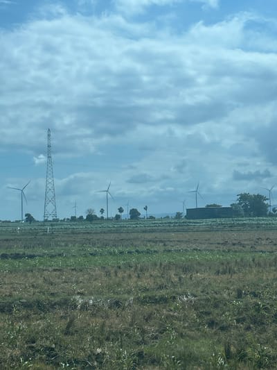 The image depicts a wide, open field with agricultural land in the foreground. In the mid-ground, there are several wind turbines, indicating a wind farm. A tall transmission tower stands prominently among the turbines, suggesting the generation and distribution of electricity. The background reveals a cloudy sky with a light blue hue, hinting at potentially overcast weather. The scene appears to be rural, characterized by fields and the presence of renewable energy infrastructure. The image does not contain any people or specific actions or interactions.