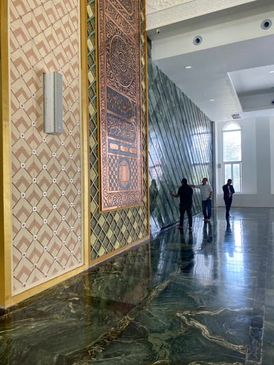 The image depicts the interior of a building with elaborate wall and floor decorations. On the left, a wall features a geometric pattern of beige and brown diamonds with small red squares at the intersections, and a vertical speaker is mounted on it. Adjacent to this is a tall, narrow panel with intricate copper-colored Islamic calligraphy and geometric designs. To the right, a slanted wall is covered in reflective green marble tiles.

Three men are present in the scene. The first man, on the far left, is wearing a black shirt and dark pants, facing the marble wall and touching it with his left hand. The second man, in the center, is wearing a white shirt and blue jeans, with his right hand resting on the marble wall and his head tilted as if in conversation with the first man. The third man, on the far right, is wearing a dark suit jacket and pants, standing with his hands clasped in front of him and looking towards the right side of the frame.

The floor is made of highly polished dark green marble with swirling lighter veins, reflecting the light from the ceiling and the windows. In the background, a tall, arched window with decorative grilles is visible, letting in natural light. The ceiling is white, with recessed lighting and a decorative plaster border. The overall impression is one of an opulent and architecturally significant space, likely a place of worship or a cultural center. The location is identified as Aluekambuk, Indonesia, suggesting it could be a mosque or a building of similar importance within that community.