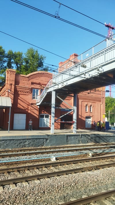 A brick building at a train station in Vyshny Volochyok, Russia, is visible under a clear blue sky. The building is painted red and has a white door and several windows. A pedestrian bridge crosses over the train tracks, with stairs leading up to it. There are several people standing on the platform near the bridge, some wearing yellow vests. Train tracks are in the foreground, with ballast and gravel.