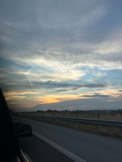 The image captures a view from a car window, looking out at a road and a sunset. The road is paved with asphalt and has a white line marking the lane. A metal guardrail runs along the side of the road, separating it from a dry, grassy field. In the distance, a line of trees and hills can be seen against the horizon. The sky is filled with dramatic clouds, tinged with orange and yellow hues from the setting sun. The overall scene suggests a journey through the countryside of Manolsko Konare, Bulgaria, during the evening. The image does not contain any people.