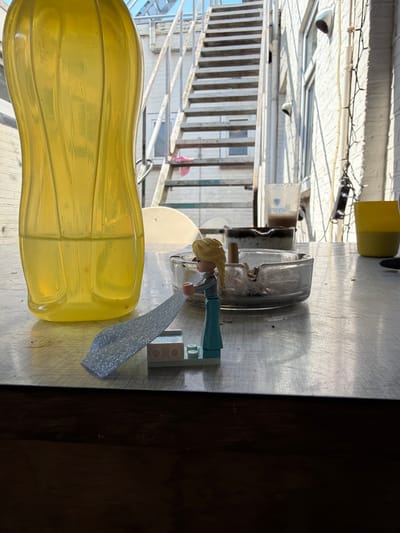 An outdoor patio scene in Boxtel, Netherlands, features a table with various objects. A large, translucent yellow bottle with vertical ridges stands on the left side of the table. In the center, a glass ashtray contains cigarette butts. A small figurine, appearing to be a Lego character with blonde hair and a blue dress, is positioned in front of the ashtray, holding a silver, textured rectangular object. A white, patterned fabric or sheet is partially draped over the figurine and extends towards the bottle. To the right of the ashtray are two cups: a clear plastic cup with a dark liquid, and a yellow cup. In the background, a metal staircase with many steps ascends alongside a brick building. The scene is illuminated by sunlight, casting shadows.