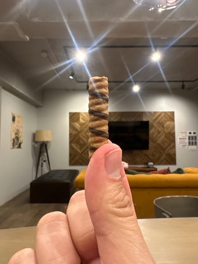 The image displays a human hand holding a ridged, cylindrical snack, positioned vertically in the foreground. The snack exhibits alternating light and dark brown spiraling stripes. The hand, with light-toned skin, grasps the object between the thumb and index finger.

The background reveals an interior common area or lounge located in Nakano, Japan. A light-colored table surface is visible at the bottom right. Dominating the midground is a mustard-yellow sofa or sectional, draped with dark green and maroon textiles. Behind the sofa, a large, dark-screened flat-screen television is mounted against a wall adorned with light brown, geometrically patterned wooden panels. To the left of the television, a light grey wall features a framed poster with a light background and dark figures, positioned above a tripod floor lamp with a beige, rectangular shade. A dark, rectangular upholstered bench or ottoman rests on the light brown wooden floor.

The light grey ceiling displays visible beams or conduits, along with several circular light fixtures emitting strong lens flares. A small, white, circular ceiling-mounted device, consistent with a smoke detector, is also present, alongside black track lighting fixtures with spotlights.