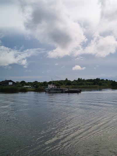 The image depicts a serene river scene in Kargulino, Russia. The foreground is dominated by the rippling surface of the water, reflecting the sky above. In the mid-ground, a ferryboat is docked, appearing to be ready for or just completing a journey. The ferry is gray and white, with a few people visible on its deck. Behind the ferry, a dense line of lush green trees forms the horizon, suggesting a natural, possibly rural, environment. To the left of the ferry, a dark-colored building with a white section on its facade is situated near the water's edge, possibly a boathouse or a small structure associated with the ferry service. The sky is a mix of dramatic clouds and patches of blue, indicating varied weather conditions. The overall impression is one of tranquility and everyday life by the river.