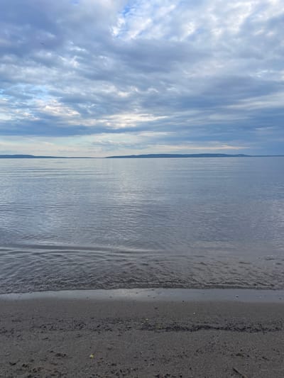 The image depicts a serene natural landscape, likely a coastline or lakeside, under a cloudy sky. The foreground features a sandy beach, with the gentle waves of a body of water lapping at the shore. In the distance, a flat horizon line suggests landmasses or hills. The sky is filled with a layer of overcast clouds, with some breaks allowing diffused light to illuminate the scene. The overall impression is one of tranquility and natural beauty, characteristic of outdoor environments. The location is identified as Primorsky, Russia, suggesting a coastal setting within this region. The scene is devoid of any human presence or man-made structures, focusing solely on the natural elements of sand, water, and sky.