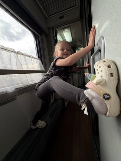 A young child, likely a girl with blonde hair, is depicted hanging upside down in a playful manner inside what appears to be a train carriage or a long-distance bus. Her left hand is gripping an overhead grey handle or fixture on the light-colored ceiling, while her left foot, clad in a white Croc shoe adorned with colorful unicorn and glitter charms, is pressed against the ceiling. Her right foot, also wearing a white Croc with a light blue strap, rests against a dark, slatted panel running along the lower wall of the vehicle.
The child is dressed in a dark patterned short-sleeved top and dark leggings, and looks directly at the camera with a neutral expression. The interior features light-colored walls, overhead lighting, and windows with white, ruffled sheer curtains. Through the windows, an overcast or cloudy sky is visible, indicating daytime. The Crocs brand logo, including the word "CROCS" and the crocodile emblem, is clearly visible on the shoes. The scene captures a lighthearted moment within a transit setting in Urusovo, Russia.