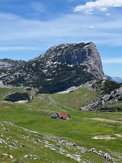 The image shows a landscape in Dubrovsko, Montenegro. The dominant feature is a large, rocky mountain with patches of snow and vegetation clinging to its slopes. To the right of the mountain, a steep, grassy hillside slopes down to a small cluster of buildings with red and grey roofs. The sky above is blue with streaks of clouds. The scene is bathed in daylight.