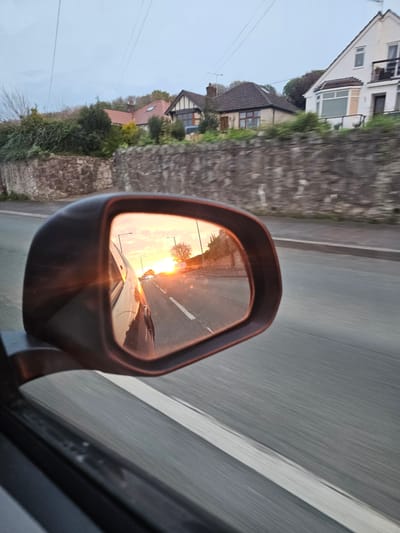 The image depicts a car side mirror reflecting a sunset on a road in Holway, United Kingdom. The mirror shows the road stretching into the distance with the sun setting at the end. In the foreground, the side of the car and the frame of the mirror are visible. The rest of the scene outside of the side mirror shows a road and a roadside view of houses.