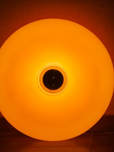 This is a close-up, top-down shot of a round, orange lamp. The lamp is illuminated from within, casting a warm, orange glow. In the center of the lamp, there is a dark, circular opening. The background is composed of dark, textured surfaces, likely walls or a ceiling, with hints of wood grain in the right side. The lighting creates a diffused, ambient atmosphere. No people, text, or specific location cues are visible to indicate the time of day, weather, or emotions. The context of Tyrnyauz, Russia is not visually represented in the image.