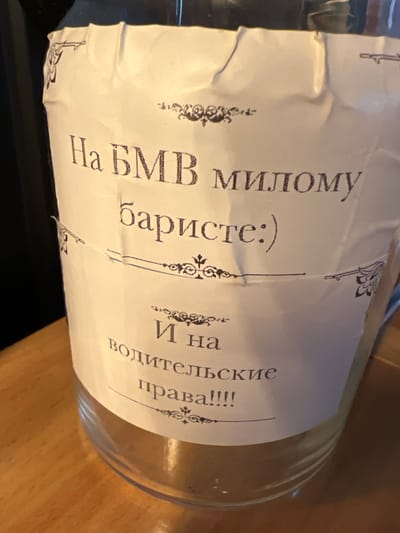 The media file shows a close-up, side-view of a glass bottle with a label that appears to be handwritten or printed in a decorative font. The bottle is positioned on a wooden surface, likely a table, and is tilted to the right, obscuring much of its contents. The lighting suggests an indoor setting, possibly in the evening.

The label is primarily white with ornate black decorative elements, resembling flourishes or small illustrations, placed at intervals. Visible text on the label is in Russian. The prominent text reads: "На ВМИВ Милому баристе :)" and "И на водительские права!!!!". The first line translates to "To the VMIV Beloved Barista :)", and the second line translates to "And for driver's licenses!!!!". The presence of the smiley face emoji suggests a lighthearted or humorous message.

The overall impression is of a gift or a personal message attached to a bottle, possibly for a barista, with an added, perhaps ironic, mention of driver's licenses. The bottle itself appears to be clear glass. The wooden surface provides a warm, textured background. The image's focus is on the label and the text, with the bottle and its surroundings serving as supporting elements.