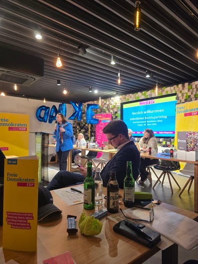 An indoor political gathering of the "Freie Demokraten FDP" (Free Democratic Party) is depicted, occurring in Hagen, Germany, on March 26, 2026, as indicated on the large projection screen.

The scene features a modern industrial-style interior with a dark, corrugated metal ceiling equipped with recessed spotlights, exposed filament bulbs, and an air conditioning unit. Walls include yellow "Freie Demokraten FDP" banners and a mosaic-patterned wall composed of wooden blocks in light, dark, and green tones. Blue foil balloons spelling a partial word (e.g., "ANKÉE") are positioned behind the speaker.

Approximately seven individuals are visible. In the midground, a woman in a blue jacket and a patterned scarf is standing at a transparent podium, holding a microphone, and gesturing while speaking to an unseen audience. Seated at wooden tables in the midground and background are several attendees. One man in a dark jacket is seated, looking down, possibly at a laptop or papers. A woman to his right is observing the speaker. Another partially visible person in the foreground is seated, holding a mobile phone; a black "BOSS" brand baseball cap rests on their table.

Tables are laden with various items: two green glass beer bottles (one labeled "Pülleken"), a clear water bottle, small Coca-Cola bottles, a black pen, eyeglasses, white papers, a black phone case or wallet, a blue "FDP-FW" branded chocolate bar wrapper, and a crumpled light green paper or napkin. A yellow "Freie Demokraten FDP" leaflet, displaying the party's slogan "Jeder Einzelne hat es in der Hand. Veränderung, Chancen, Aufbruch. Unser Auftrag ist dabei klar. Stärken wir den Glauben der Menschen an sich selbst," is prominently placed on a table in the foreground. A box with "Deutsche Krebshilfe" branding and what appear to be bottle caps on top is also visible on a table in the background.
