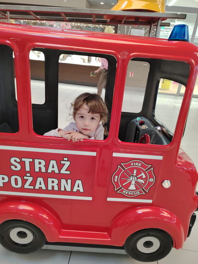 A young child with curly brown hair is sitting in a toy fire truck. The child is leaning on the edge of the truck, with their arms crossed. They are looking directly at the camera with a neutral expression. The fire truck is red and has "STRAŻ POŻARNA" written on the side, which translates to "FIRE BRIGADE" in Polish. It also features a "FIRE RESCUE" emblem. The truck has blue lights on top and a steering wheel and dashboard inside. The background shows a retail environment, possibly a shopping mall, with displays and other people visible in the distance. The floor is tiled. The setting appears to be indoors, during daylight hours.