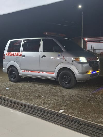 A silver minivan, modified as an ambulance, is parked on a gravel surface at night in Bulo Gogo, Indonesia. The vehicle displays "AMBULANCE" in red capital letters on its rear left door, and mirrored "AMBULANCE" text is visible on the front grille. Red and white checkered and striped decals run along the lower side panel. A red heartbeat line decal and various small stickers are present on the front left fender. An orange emergency light bar is mounted on the roof, and blue and yellow emergency lights are illuminated on the lower front bumper. The vehicle's headlights are also active. It is equipped with black steel wheels and rugged tires. In the foreground, a metal grate separates the gravel from a light-colored tiled surface. In the background, a street light on a utility pole illuminates the scene, and a building with red and white features, possibly a service station, is visible to the right. No individuals are present.