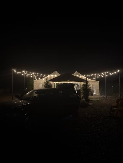 A dark, outdoor scene at night, illuminated by strings of overhead lights. In the foreground, a dark-colored car is parked, its surface glistening with raindrops. Behind the car, a white tent structure, also lit from within and around by the string lights, is visible. The ground is covered in gravel. To the right, two wooden chairs are partially visible. The overall impression is of a temporary outdoor gathering space set up at night, possibly during or after rain. The image does not provide any information about the city of Kalinkovo, Russia.