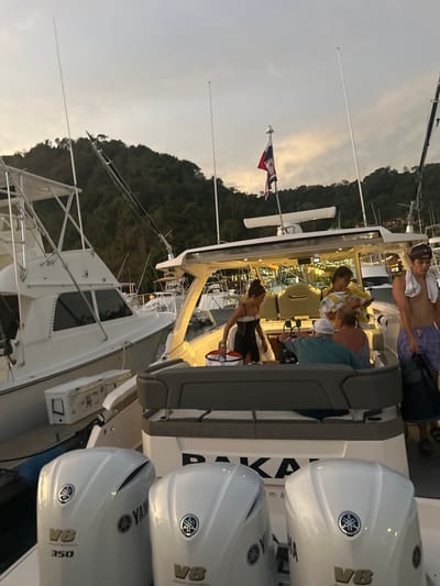 The image shows a white motorboat docked at a marina in Playa Herradura, Costa Rica. The boat is a large, luxurious vessel, with a spacious deck and a hardtop. There are several people on board the boat, some are standing near the rear and some are seated inside the cabin. One woman in a black dress is standing on the deck, while another woman is seated in a cabin chair. There are three outboard motors attached to the rear of the boat, all of which are white and have Yamaha branding. The boat is flying a Costa Rican flag on its stern. The boat is docked alongside another larger white boat, which is also docked at the marina. The background is made up of lush green vegetation and a blue sky. The image was taken at dusk, as the sun is setting in the background.