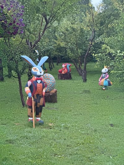 In a grassy park area surrounded by trees, a life-sized statue of a blue rabbit wearing a colorful suit and holding a cane stands prominently in the foreground. Behind the rabbit, a large, patterned egg sculpture rests on a tree stump. To the right, a smaller statue of a rabbit wearing a crown and a colorful dress is visible. Further in the background, a statue of a squirrel is perched on a tree stump. The scene is set in the city of Kalinkovo, Russia.