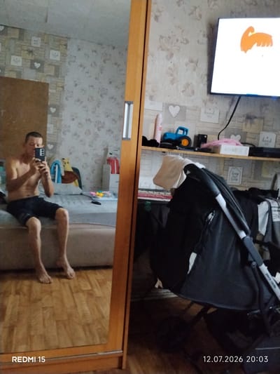 A man, shirtless and wearing dark shorts, is captured taking a mirror selfie in what appears to be a bedroom. He is sitting on the edge of a bed with a light-colored duvet. The room has patterned wallpaper with floral motifs and heart shapes. A wooden closet with a mirror door frames the scene on the right. In the reflection of the mirror, a television screen displays an animated orange graphic, and a blue toy car is visible on a shelf. There is also a baby stroller in the foreground of the reflection, partially obscuring the view of the room behind it. The floor is made of wooden planks. The bottom of the image shows the text "REDMI 15" and the date "12.07.2026 20:03". The location is not specified within the image itself.