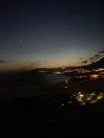 The image depicts a dark coastal scene, likely at dusk or night, in Plat, Croatia. On the left, the sky transitions from dark blue to a band of orange near the horizon. A small, faint light is visible in the darker sky. The right side of the image is predominantly dark, but there are streaks of yellow and orange lights, likely from a town or city on the coast. These lights are blurry and appear to reflect in water. The image seems to be taken from a higher vantage point, possibly a hillside or building overlooking the coastline.