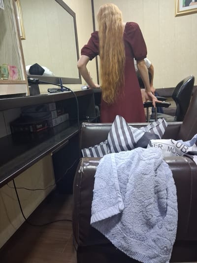 The image shows a woman with long, blonde hair standing in what appears to be a salon or beauty shop. She is wearing a maroon dress. She is holding her hair, possibly preparing to cut it or style it. In the background, a brown leather chair is visible, with a light blue towel draped over it. There are also some striped fabrics placed on the chair. A mirror is on the left side of the image, reflecting part of the room, including some shelves with various items. Below the shelves, there is a drawer filled with boxes, some of which have red labels. The overall setting seems to be indoors, with warm lighting. Some items are stacked on shelves, including what appears to be cash or vouchers. There is visible text on some of the boxes, including "Geeany". The image is captured in Kahramon, Uzbekistan.