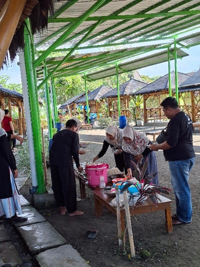 The image depicts a group of people gathered under a green awning in Kecamatan Tulung, Indonesia (latitude: -7.592, longitude: 110.65). Five individuals are present, four women and one man. Two women are seated at a table, with a woman standing beside them and a man standing behind them. Another woman is standing away from the table. There are two wooden tables with a pink cooler, food, and various containers on them. The group appears to be engaged in a meal. The surrounding environment includes a row of bamboo huts and lush greenery.