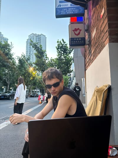 The image, taken in Putuo District, China, captures a vibrant street scene during the golden hour of a clear day, indicated by the warm, low-angle sunlight. The central subject is a young man with dark hair and sunglasses, wearing a black sleeveless top, who looks directly towards the camera. He is seated outdoors, with a black laptop (likely a MacBook) open in front of him, and he holds a lit cigarette in his right hand. He also wears rings on his fingers.

In the background, a woman in a white shirt and dark pants walks along the street, past parked cars and trees. Tall residential or commercial buildings rise into the clear blue sky, framed by green foliage.

Mounted on a brick wall to the left of the main subject are several signs with Chinese text. One prominent sign features red and blue lights, and the characters "平安屋" (Ping'an Wu, likely a safety station) and "长寿路派出所" (Changshou Road Police Station), along with a phone number: "电话:22048085". Another blue sign above it displays "畅杰交通科技" (Changjie Transportation Technology) and "CJ TFCTECH", advertising "【包月、错峰咨询】" (monthly, off-peak consultation) with phone numbers "137-6163-5331" and "400-6689-197". The scene portrays a casual, urban setting, with the man seemingly relaxing at an outdoor establishment.