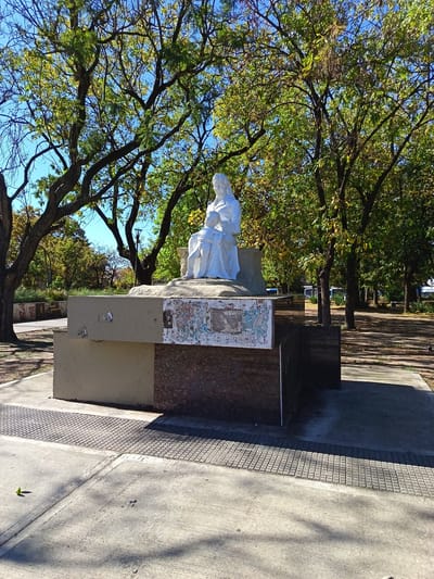 The image displays a white statue of a woman and child, likely a mother figure embracing a small child, set outdoors in a park or public square. The statue is positioned on a multi-tiered base constructed from concrete and a darker, possibly granite or marble, material. The concrete sections of the base show visible wear and tear, with remnants of faded posters, stickers, or graffiti adhering to their surfaces, particularly on the front and left side.
The scene is illuminated by bright sunlight, casting distinct shadows on the ground and indicating a clear day. The background is dominated by numerous trees with abundant green and some yellowish-green foliage, suggesting a season like late spring, summer, or early autumn. A vibrant blue sky is visible through the branches.
In the foreground, the ground consists of concrete paving, with a lone green leaf resting on its surface. A paved pathway is discernible winding through the trees in the background, and distant objects such as light poles and what appear to be white vehicles can be faintly seen beyond the trees on the right. The overall setting is a public green space in Remedios de Escalada, Argentina, featuring a weathered but prominent monument. No specific legible text is clearly discernible on the statue's base due to the state of the applied materials.