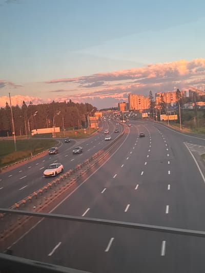 The image, rotated 90 degrees counter-clockwise, presents an elevated view of a multi-lane highway or motorway during what appears to be sunset or sunrise. The scene captures a moment during the "golden hour," bathing the landscape in warm, orange-pink light.
The primary subject is the expansive highway, curving gently through the frame. Multiple vehicles, including cars and at least one truck, are visible on the road, indicating active traffic flow in both directions. Lane markings, consisting of dashed white lines, are clearly visible, as are solid white lines and a concrete barrier separating the main thoroughfare. A red circular "No Entry" sign is visible on the median strip.
To the left of the highway, a series of multi-story residential buildings with orange facades are illuminated by the low sun, creating a striking contrast with the blue sky above. Below these buildings, trees and streetlights line the road. To the right of the highway, a dense forest or wooded area runs parallel to the road.
The sky above is a notable feature, displaying a gradient from a clear, soft blue at the top to vibrant orange and pink hues where scattered clouds catch the last or first rays of the sun. The perspective is from an elevated vantage point, likely a bridge or overpass, with a part of its structure visible in the foreground. No people are discernible in the image. The overall impression is one of calm and tranquility, despite the movement of traffic. The location context is посёлок опытного хозяйства «Ермолино», Russia.