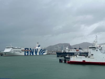 The image displays a bustling commercial port scene under an overcast sky in Ksar El Majaz, Morocco. The foreground features calm, greenish-blue water with a white and red vessel, identified as "AL ANDALUS EXPRESS," positioned on the right, its bow facing right, partially obscuring its full length. In the mid-ground, a large white ferry named "EXCELLENT" with prominent blue "GNV" branding is visible on the left, seemingly docked. Behind and to the right of the "AL ANDALUS EXPRESS" is another dark-hulled vessel bearing "DFDS" markings. A smaller black and white tugboat is visible between these ships. The background reveals a coastline featuring multiple large gantry cranes, indicative of a container terminal, against a backdrop of distant, hazy mountains. No discernible human figures are visible. All vessels appear stationary.