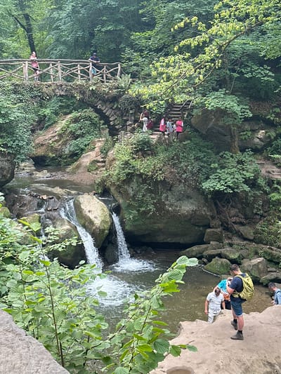 The image depicts a scene in the Mullerthal region of Luxembourg, likely near Consdorf, featuring a picturesque waterfall and a rustic wooden bridge. In the foreground, lush green foliage frames a natural environment with rocky outcroppings and a body of water. A small waterfall cascades over moss-covered rocks into a pool where three individuals are present. One person, wearing a light-colored shirt and pants, is stepping into the water. Another, a young man with a yellow backpack, is standing on a rock, taking a photo with his phone. A third individual, partially visible, is also near the water's edge.

Further back, the wooden bridge spans over a rocky section, with two individuals standing on it. The bridge is adorned with interwoven branches and leads to a stone archway. A set of wooden stairs is visible on the right side, ascending the rocky embankment. Several other people, including children wearing bright pink shirts, are gathered near the top of the stairs, appearing to be observing the scenery. The environment is characterized by dense green vegetation, including trees and ivy covering the rocks, indicating a woodland setting. The lighting suggests it is daytime, with diffused light filtering through the trees.